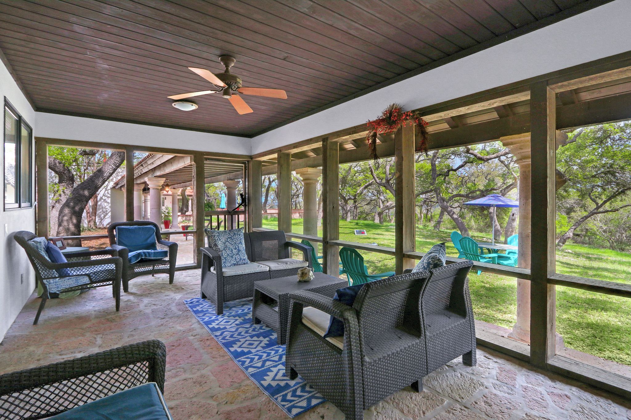 A covered patio area with wicker furniture, a ceiling fan, and large windows overlooking a grassy area.
