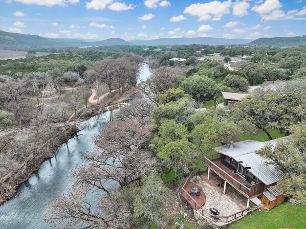 Propiedad junto al río con muelle de madera, mobiliario exterior y vistas al río y al paisaje circundante.