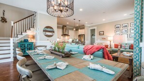 A spacious living room with a wooden dining table set for four, a teal sofa, and a staircase leading to the upper floor.