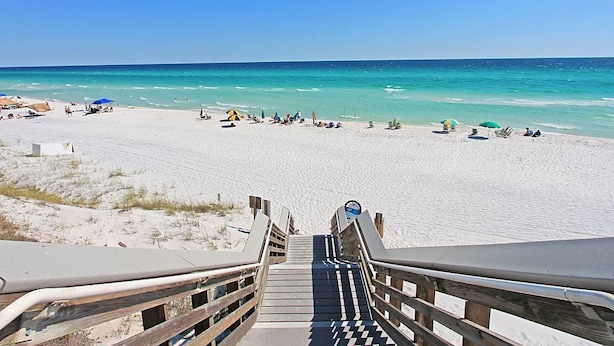 A beach with a wooden staircase leading down to the sand, a clear blue sky, and a calm sea.