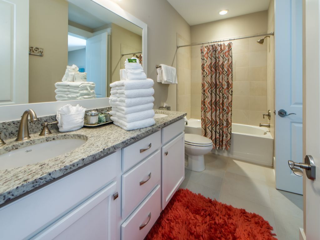 A bathroom with a sink, mirror, stacked towels, toilet, shower, and a red rug.