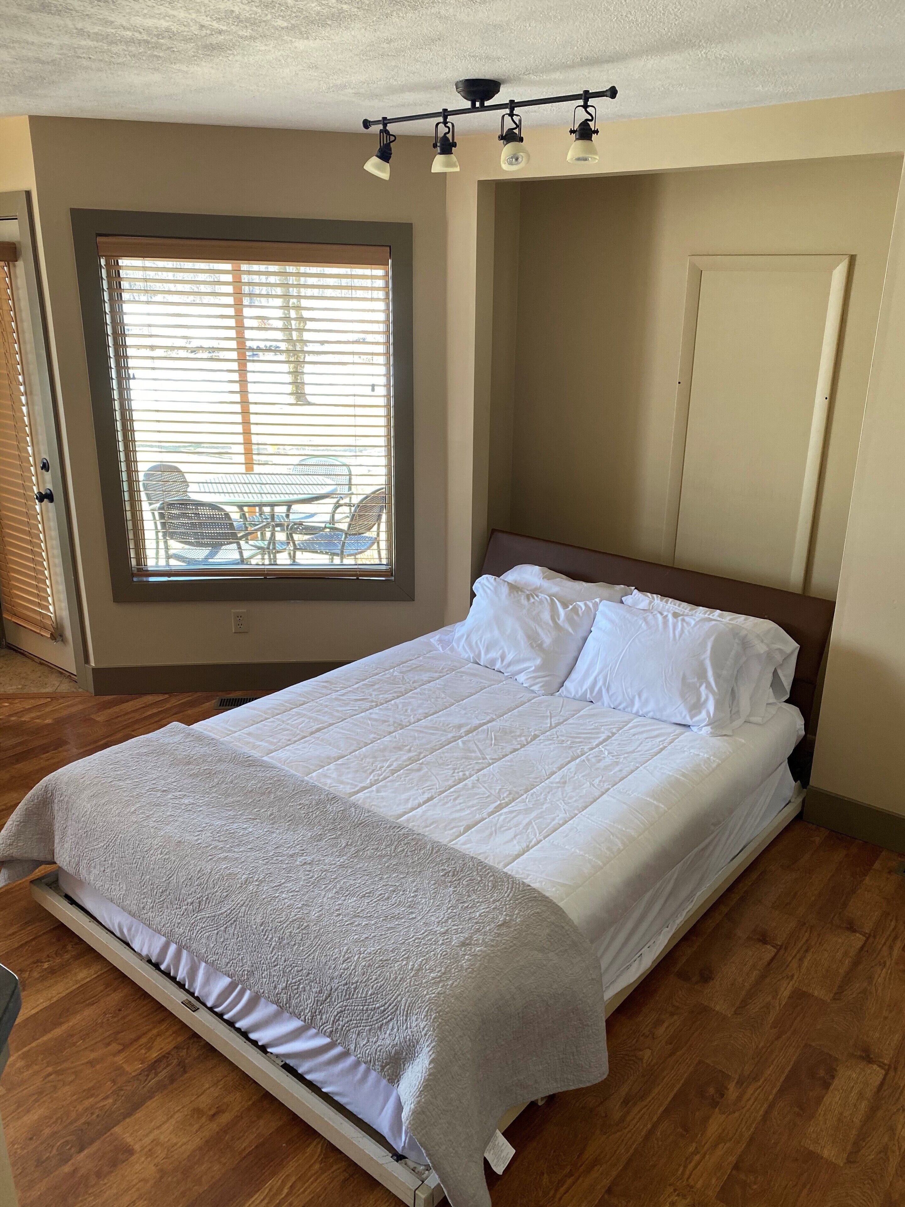 A neatly made bed with white linens in a room with a large window, a ceiling light fixture, and a door.