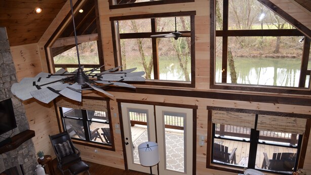 A spacious room with a large wooden ceiling, a fireplace, a dining area, and large windows overlooking a body of water. - Riverfront cabin on the Toccoa River w/hot tub  NO HIDDEN FEES (Mineral Bluff)