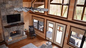 A spacious living room with a stone fireplace, wooden floors, a ceiling fan, and large windows offering views of the outdoors. - Riverfront cabin on the Toccoa River w/hot tub  NO HIDDEN FEES (Mineral Bluff)