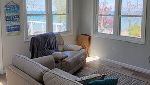 Un salon avec un canapé d’angle beige, une petite table et un tapis. Il y a deux fenêtres qui donnent sur des arbres et un plan d’eau.