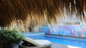 A hot tub under a thatched roof, a wooden deck, and a pool with a colorful mural.