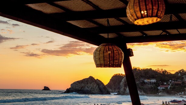 A beach view with a sunset, rocky formations, and a distant building.
