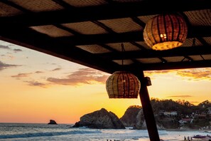 A beach view with a sunset, rocky formations, and a distant building. - Hotel Playa Zipolite (San Pedro Pochutla)