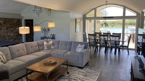 Uma sala de estar ampla com um grande sofá seccional, uma mesa de centro de madeira e uma área de refeições com cadeiras e uma mesa. Há uma parede de pedra e vista para um lago através de grandes janelas.