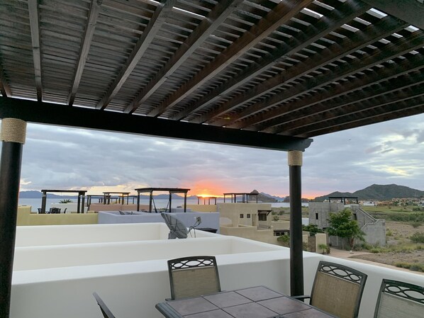 A covered outdoor seating area with a view of the sunset over a body of water and mountains.