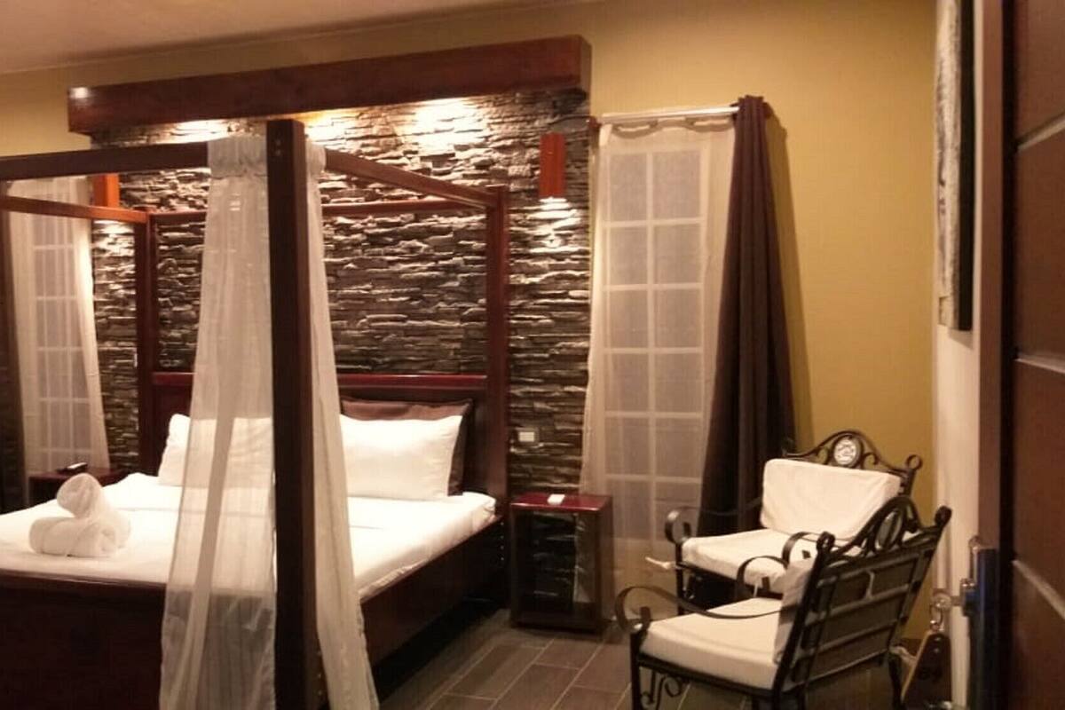 A four-poster bed with a stone wall backdrop, a bedside table, and a chair.