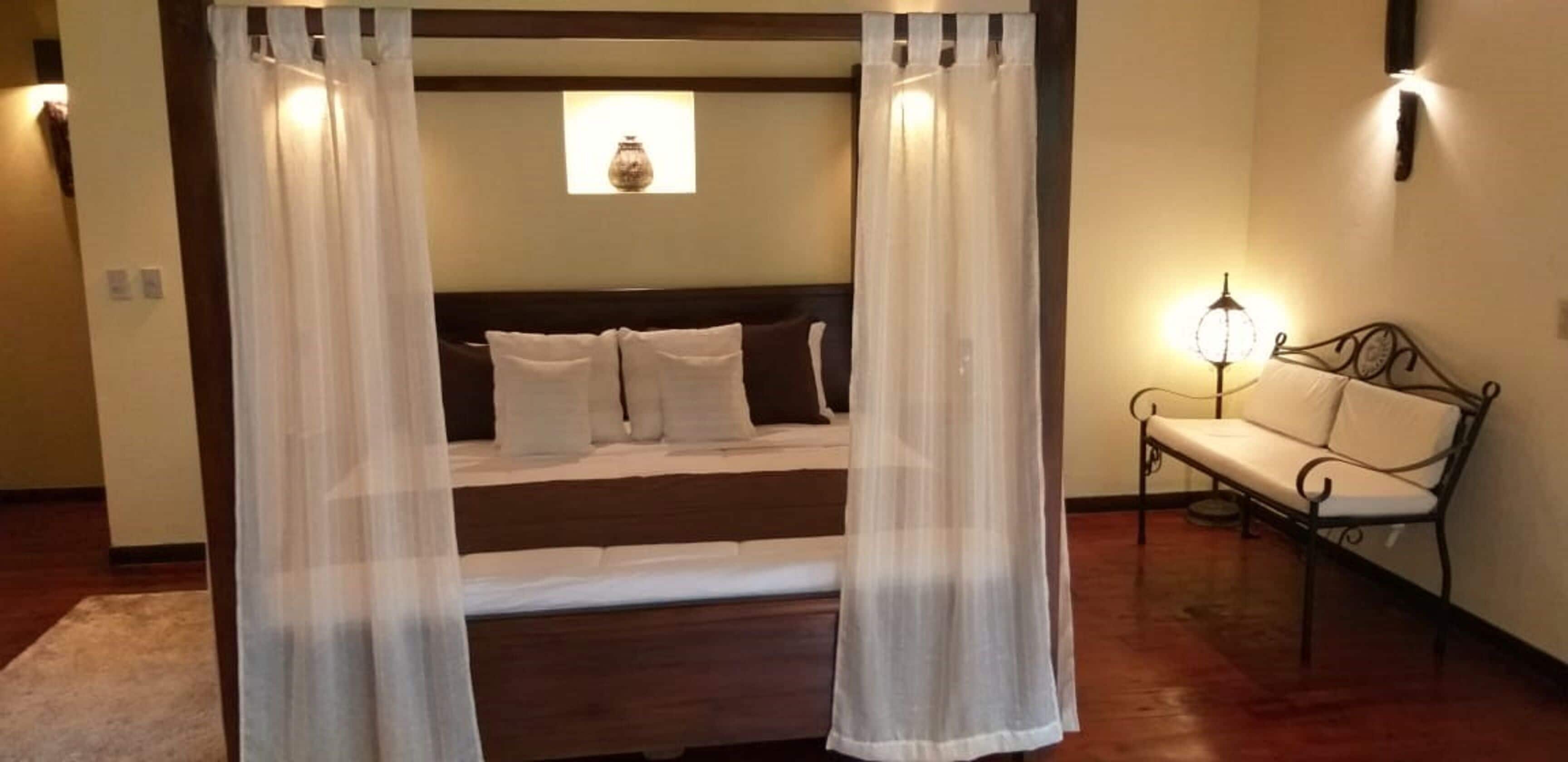 A bedroom with a bed, white curtains, a bedside lamp, and a bench.