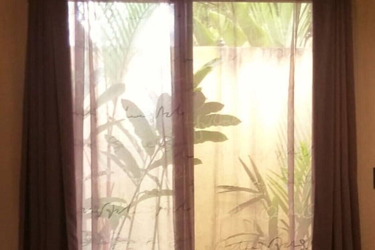 A bed with a rolled towel in front, a window with sheer curtains, and a view of greenery outside.