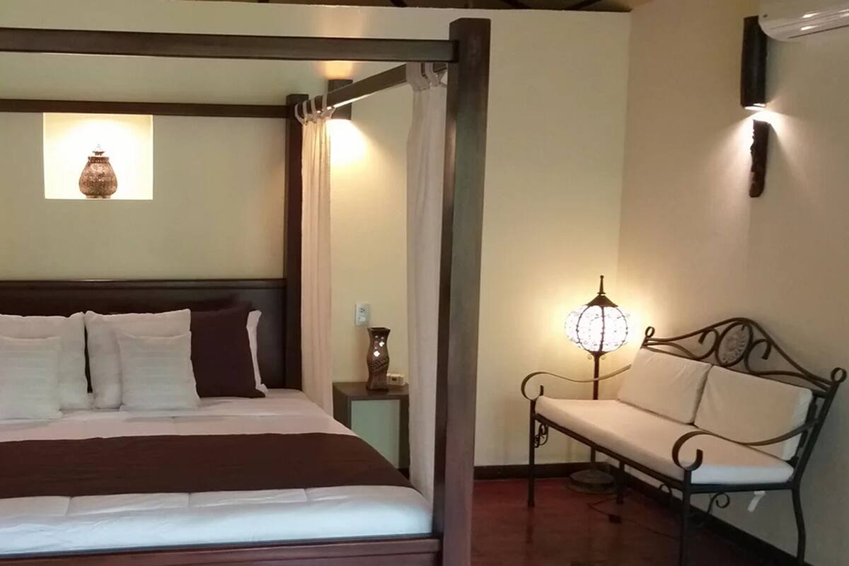 A four-poster bed with a wooden headboard, a bedside table with a lamp, and a bench with a cushion.