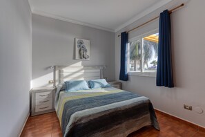 A bedroom with a bed, nightstands, a window with curtains, a wall-mounted artwork, and a wooden floor. - Gran Canaria Stays - Holiday Rentals (Mogán)