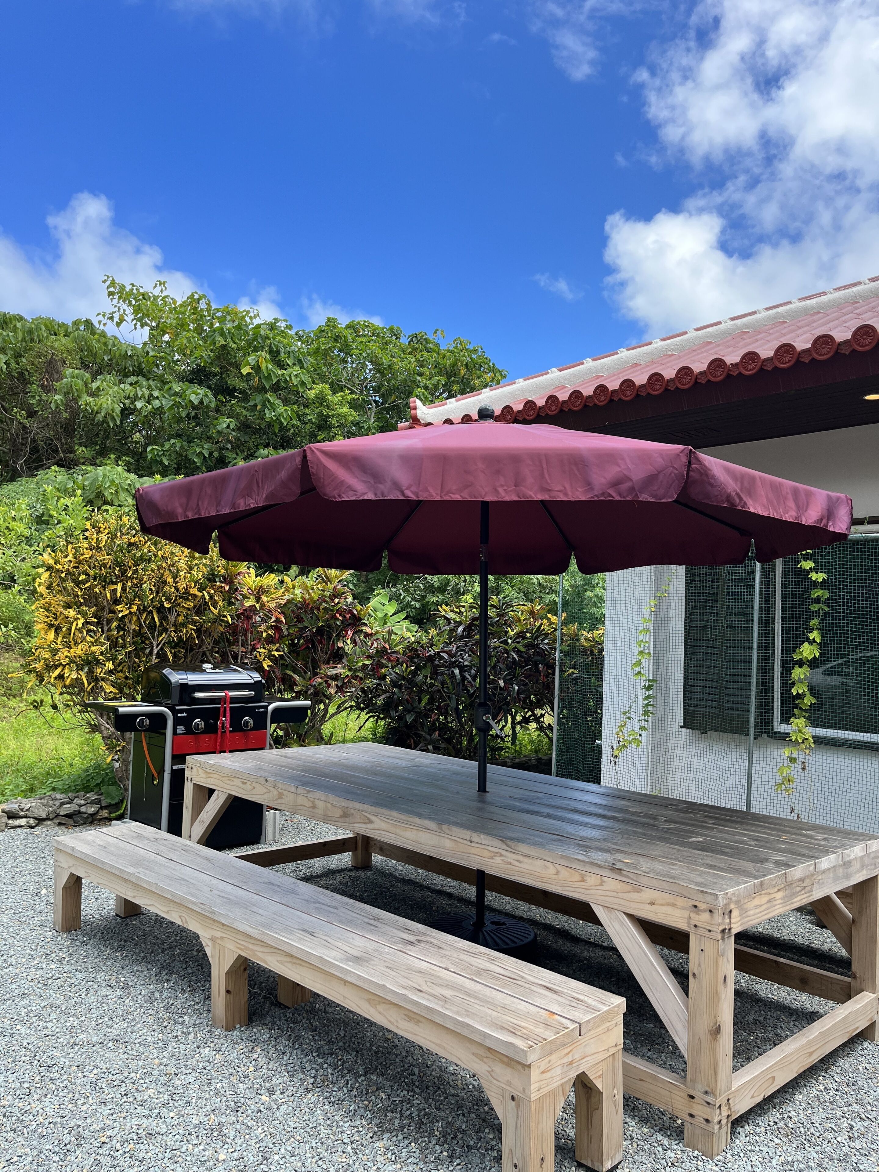 Una mesa de picnic de madera con un paraguas rojo, una parrilla y una casa con techo rojo al fondo.