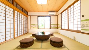 Una habitación con tatamis, una mesa redonda y cojines, puertas correderas de madera y vistas a la vegetación exterior.