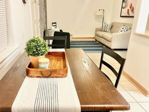 Un coin repas avec une table en bois, des chaises, un plateau sur lequel se trouvent une plante et un pot, et un canapé à l’arrière-plan.