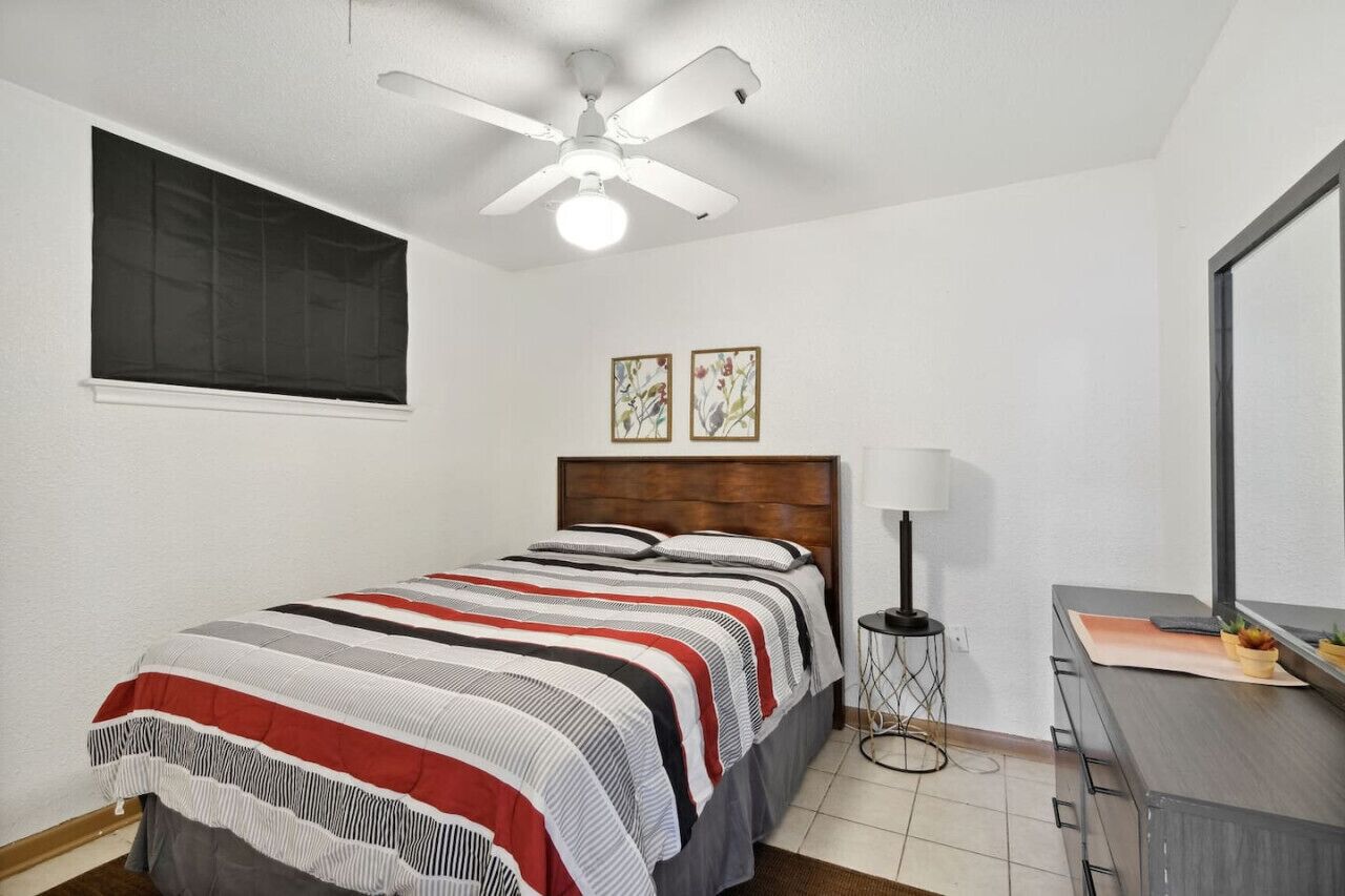 Ein Schlafzimmer mit einem Bett, einem Deckenventilator, einem Nachttisch, einer Lampe, einer Kommode und zwei gerahmten Bildern an der Wand.