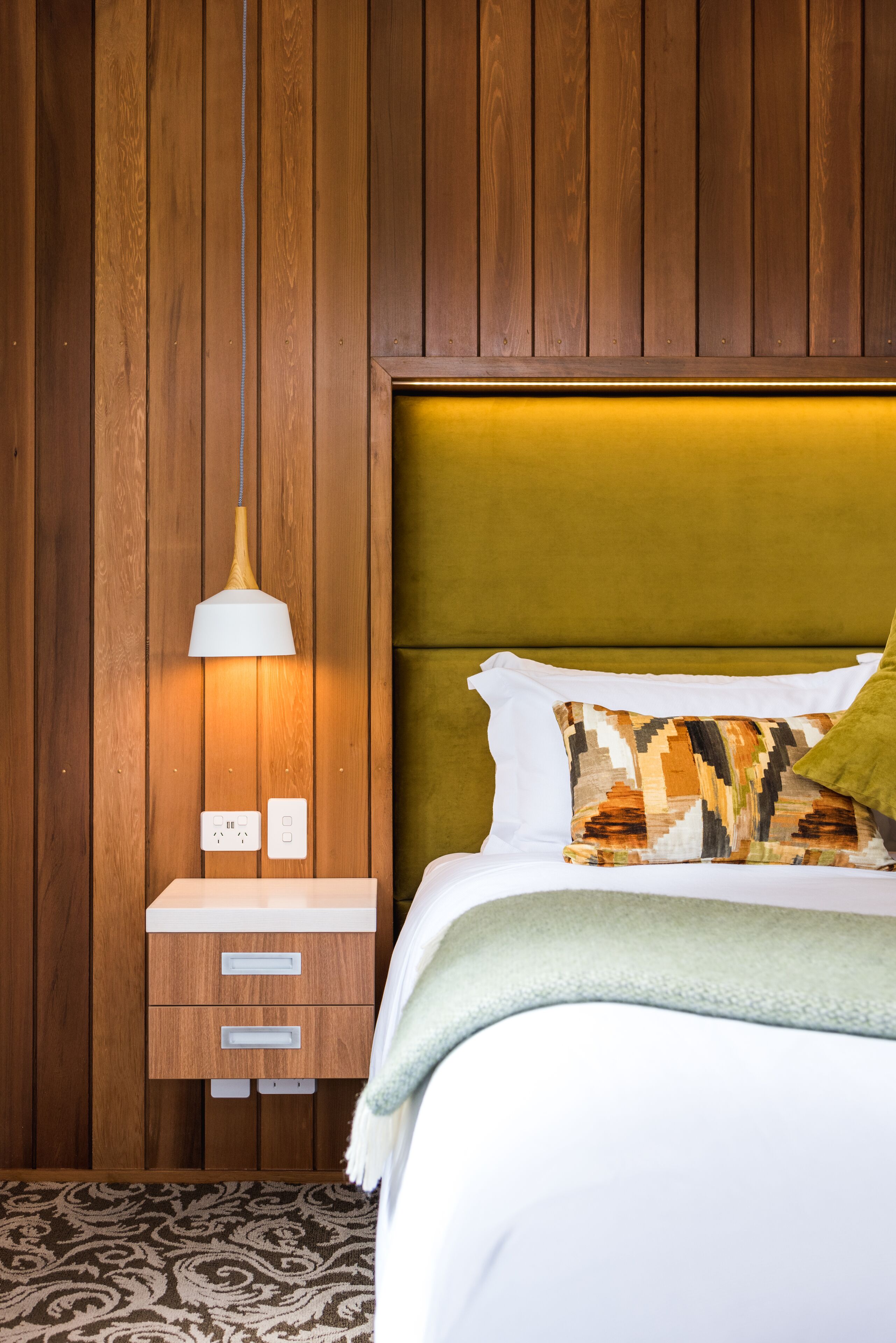 A bedroom with a wooden wall, a bedside table with drawers, a lamp, a bed with a patterned pillow, and a green throw blanket.