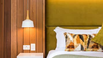 A bedroom with a wooden wall, a bedside table with drawers, a lamp, a bed with a patterned pillow, and a green throw blanket.
