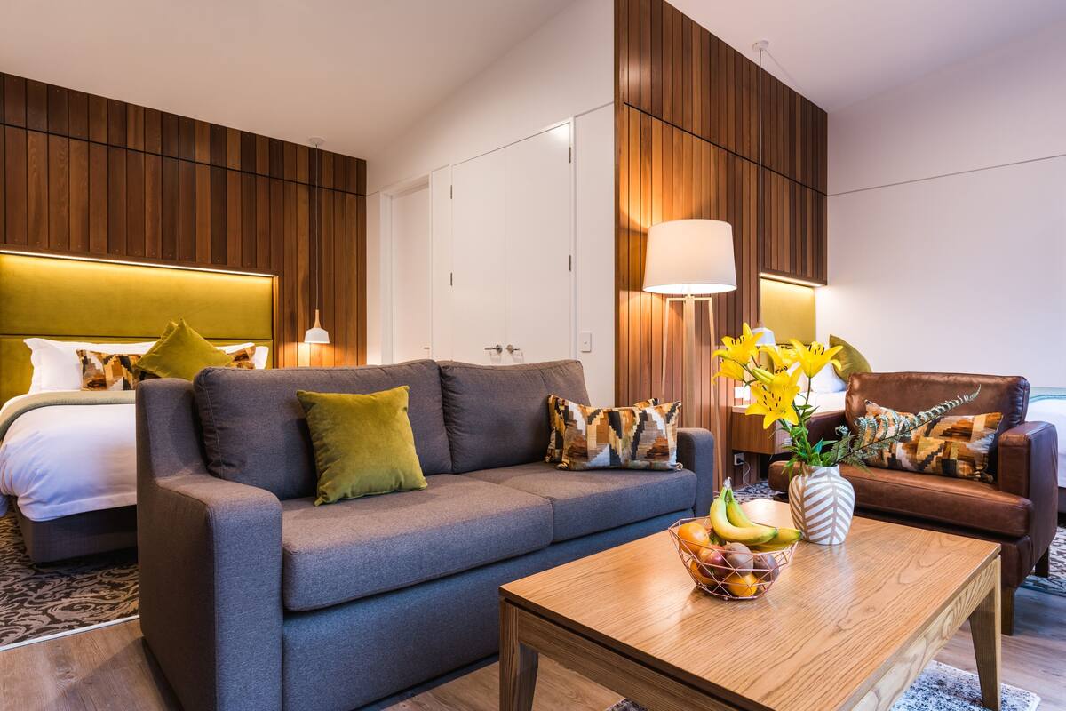 A modern hotel room with a grey sofa, a wooden coffee table with fruit, a bed with a white headboard, and a brown leather armchair.