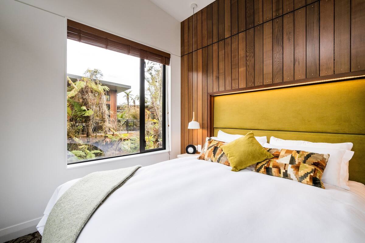 A bedroom with a large window, wooden panel wall, and a bed with multiple pillows.