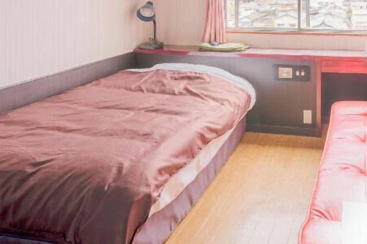 Un dormitorio con cama, escritorio con lámpara, ventana con vista a edificios y montañas, y un sofá rojo.