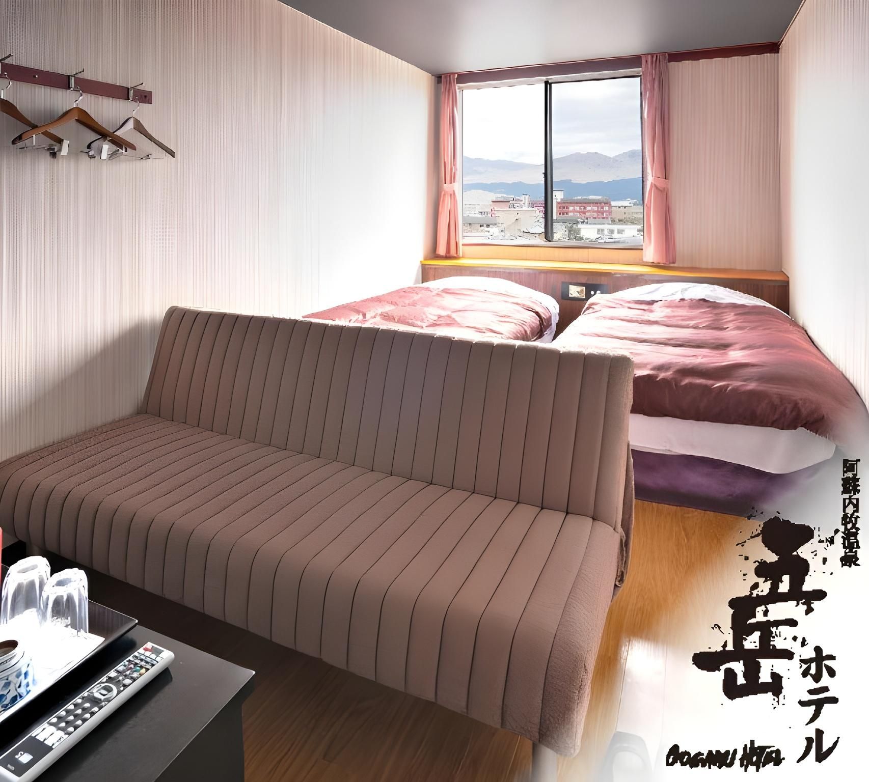 Un dormitorio con sofá, cama con ropa de cama rosa, ventana con cortinas y vistas a edificios y montañas.