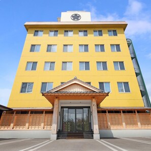 A multi-story yellow building with a central entrance, a clock on the top, and a green structure to the right. - Aso Uchinomaki Onsen Gogaku Hotel (Aso)