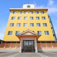 A multi-story yellow building with a central entrance, a clock on the top, and a green structure to the right.