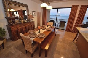 A dining area with a wooden table set for four, surrounded by wicker chairs. A large mirror and a cabinet with decorative items are on the wall.