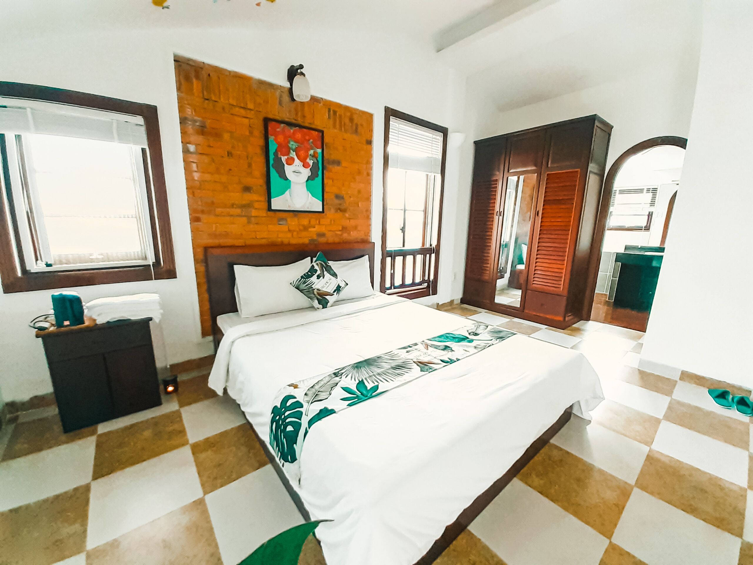 Un dormitorio con piso a cuadros, cama con colcha blanca y verde estampada, cabecera de madera y un cuadro sobre la cama.