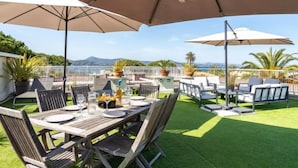 A patio with a wooden table set for dining, surrounded by chairs and large umbrellas, overlooking a scenic view with mountains and a body of water. - Les Patios by Interhome (Saint-Tropez)