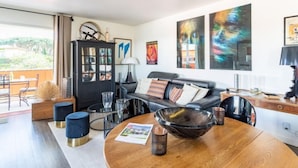 A modern living room with a black leather sofa, a glass coffee table, and a wooden dining table. There are abstract paintings on the wall and a view of the outdoors through large windows. - Les Patios by Interhome (Saint-Tropez)