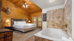 A log cabin bedroom with a large bed, a wooden chest, a ceiling fan, and a bathroom with a bathtub. - Romantic Hideaway Walk-in Cabin for Two - Fireplace & Whirlpool Tub (Branson)