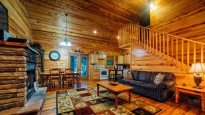 Interior acogedor de una cabaña de troncos con chimenea, piso de madera, comedor, sala de estar con sofá e incluso una escalera.