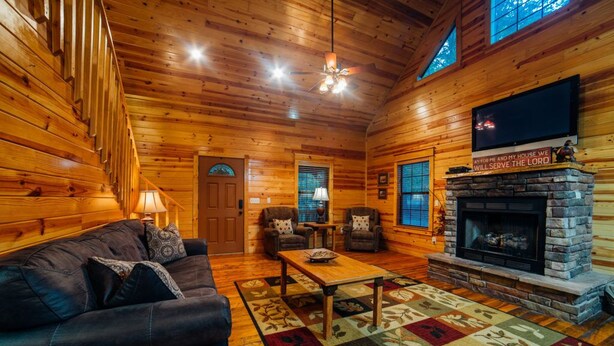 Un acogedor interior de cabaña de troncos con una escalera de madera, una chimenea, un televisor montado encima y una zona de estar cómoda con un sofá y sillones.