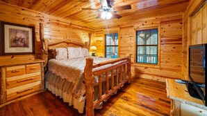 Dormitorio de cabaña de madera con una cama, un ventilador de techo, un televisor y una ventana.
