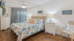 Ein Schlafzimmer mit Bett, Nachttisch, Stuhl, Kommode, Deckenventilator und einer Wanddekoration mit Strandmotiv.