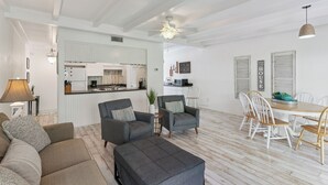 Ein geräumiger Wohnbereich mit Sofa, Hocker und Esstisch mit Stühlen. Im Hintergrund ist eine Küche erkennbar.