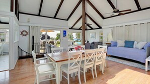 Un amplio salón con techo alto, vigas de madera, una mesa de comedor con sillas, un sofá y vistas al exterior.
