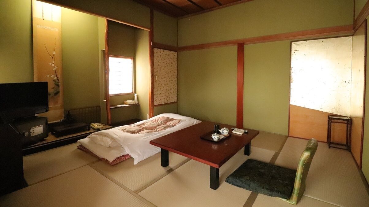 Ein traditioneller japanischer Raum mit Tatami-Boden, niedrigem Tisch, Futon und Fernseher, sowie einer Schiebetür.