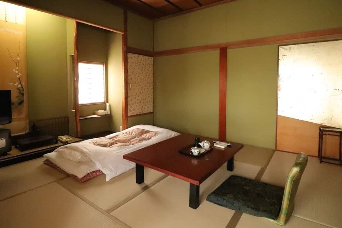 A traditional Japanese room with tatami flooring, a low table, a futon bed, a television, and a sliding door.