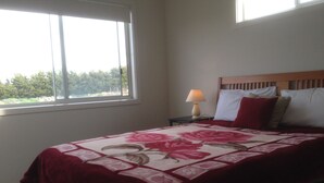 A bedroom with a large window, a bed with a floral bedspread, a nightstand with a lamp, and a wooden headboard. - Cottage Down the Lane Rural Setting (Meredith)