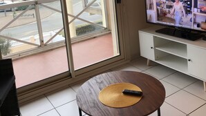 Un salon équipé d’un téléviseur, d’une porte-fenêtre coulissante donnant sur un balcon et d’une petite table ronde sur laquelle se trouve une télécommande.
