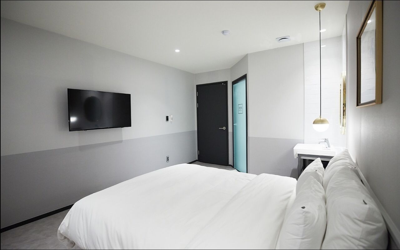 A modern hotel room with a large bed, a flat-screen TV mounted on the wall, a bedside table with a lamp, and a door leading to another room.