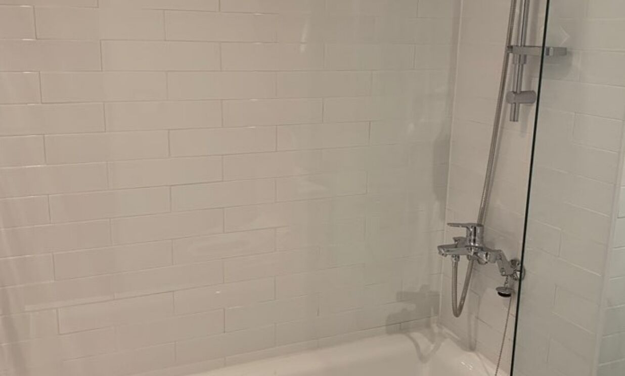 Un área de ducha con paredes de azulejos blancos y un cabezal de ducha visible, todo ello envuelto en vidrio.
