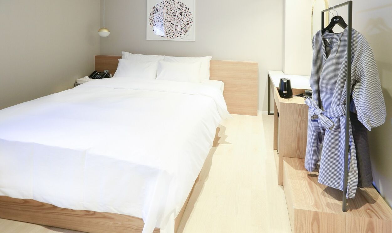 Una cama bien hecha con sábanas blancas, un cabecero de madera, una mesita de noche con un teléfono y una lámpara, y una bata colgada en un perchero.