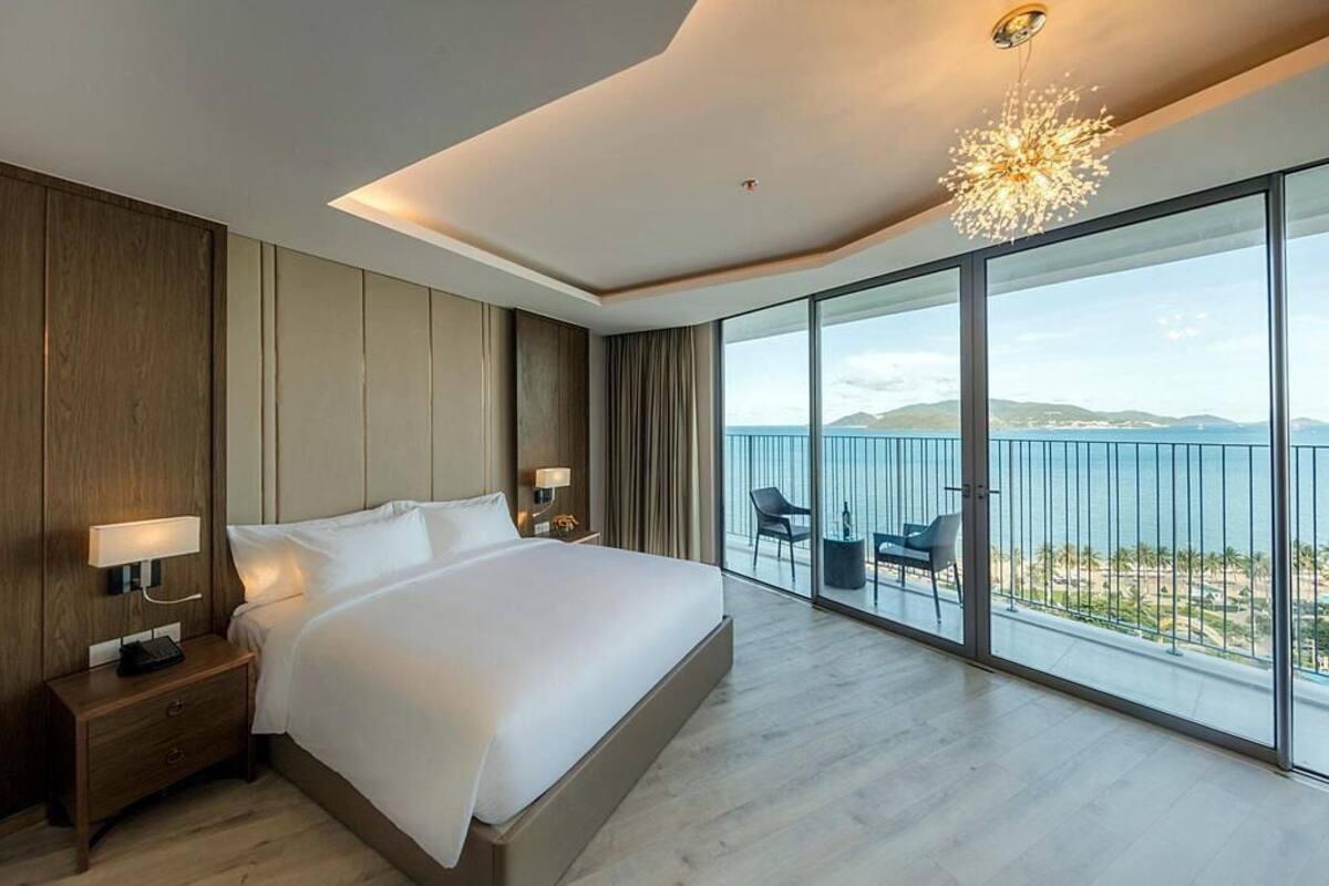 A modern hotel room with a large bed, a nightstand, a desk, and a balcony with a view of the sea.